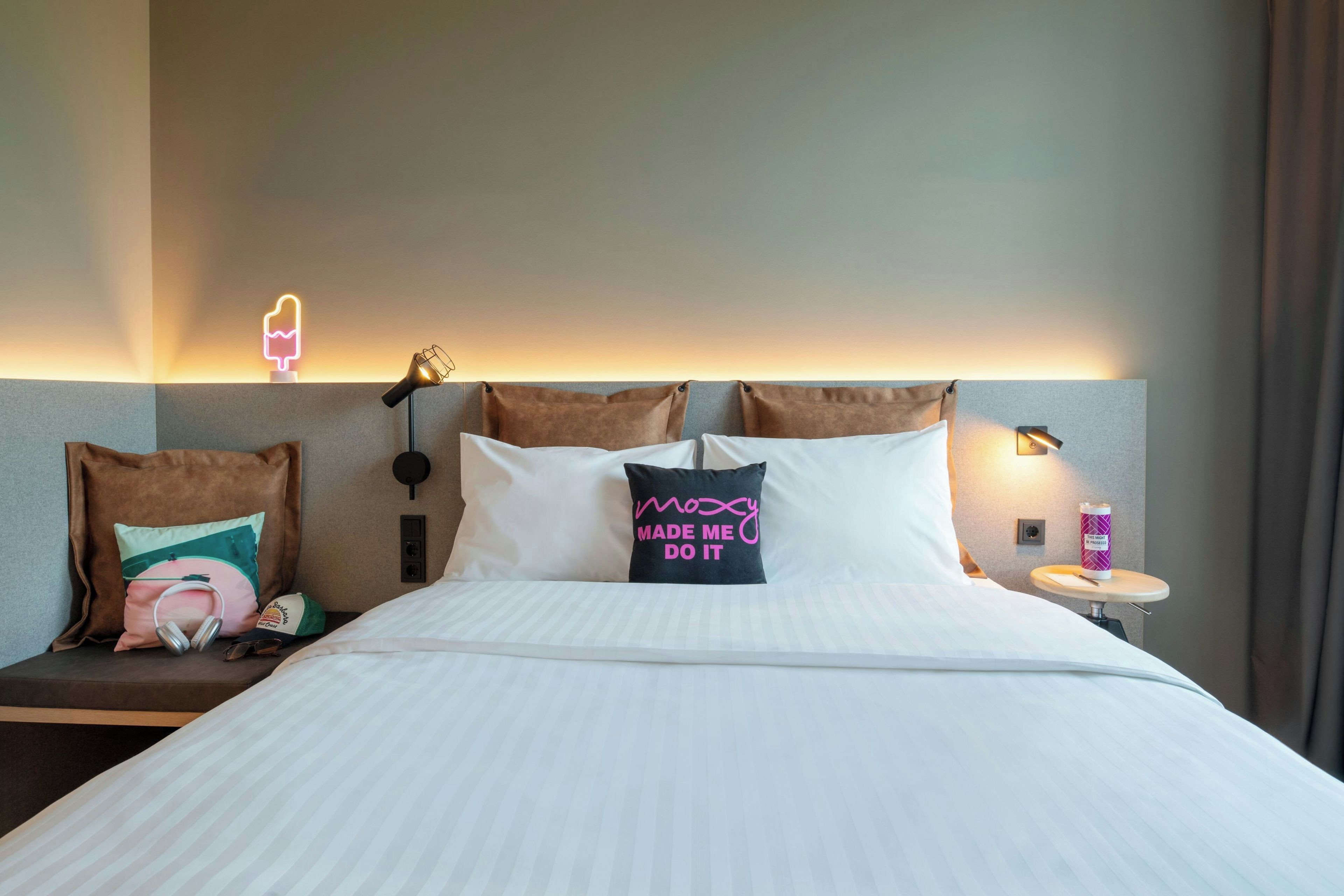 A hotel room with a bed, pillows, a nightstand, a lamp, and a neon sign.