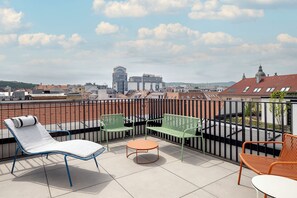 A rooftop terrace with a view of the city, featuring a white lounge chair, a green sofa, and an orange chair. - Moxy Budapest Downtown (Budapest)