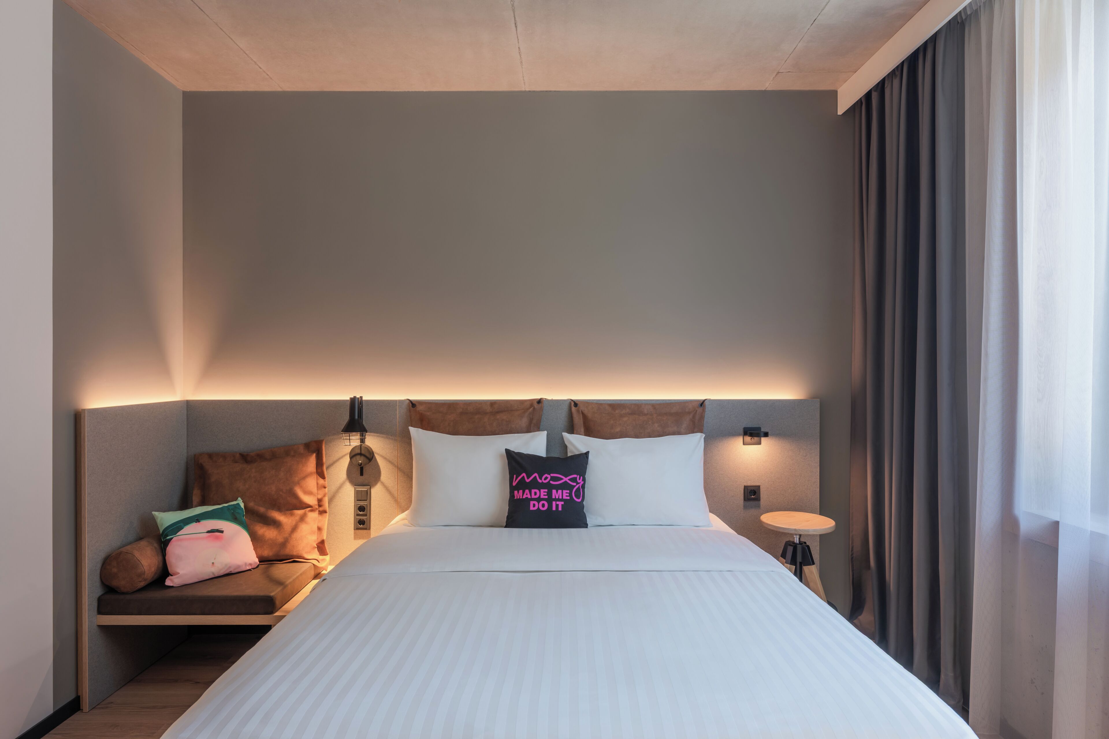 A hotel room with a bed, pillows, a bedside table, and a window with curtains.
