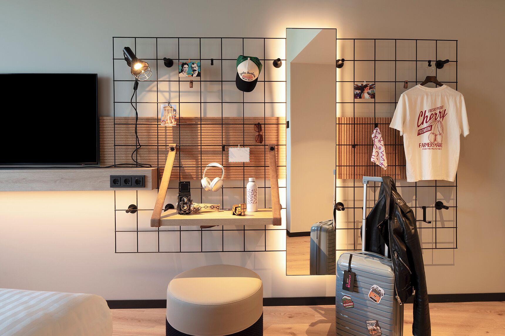 A modern hotel room with a TV, a grid wall with various items, a mirror, a bed, and a suitcase.