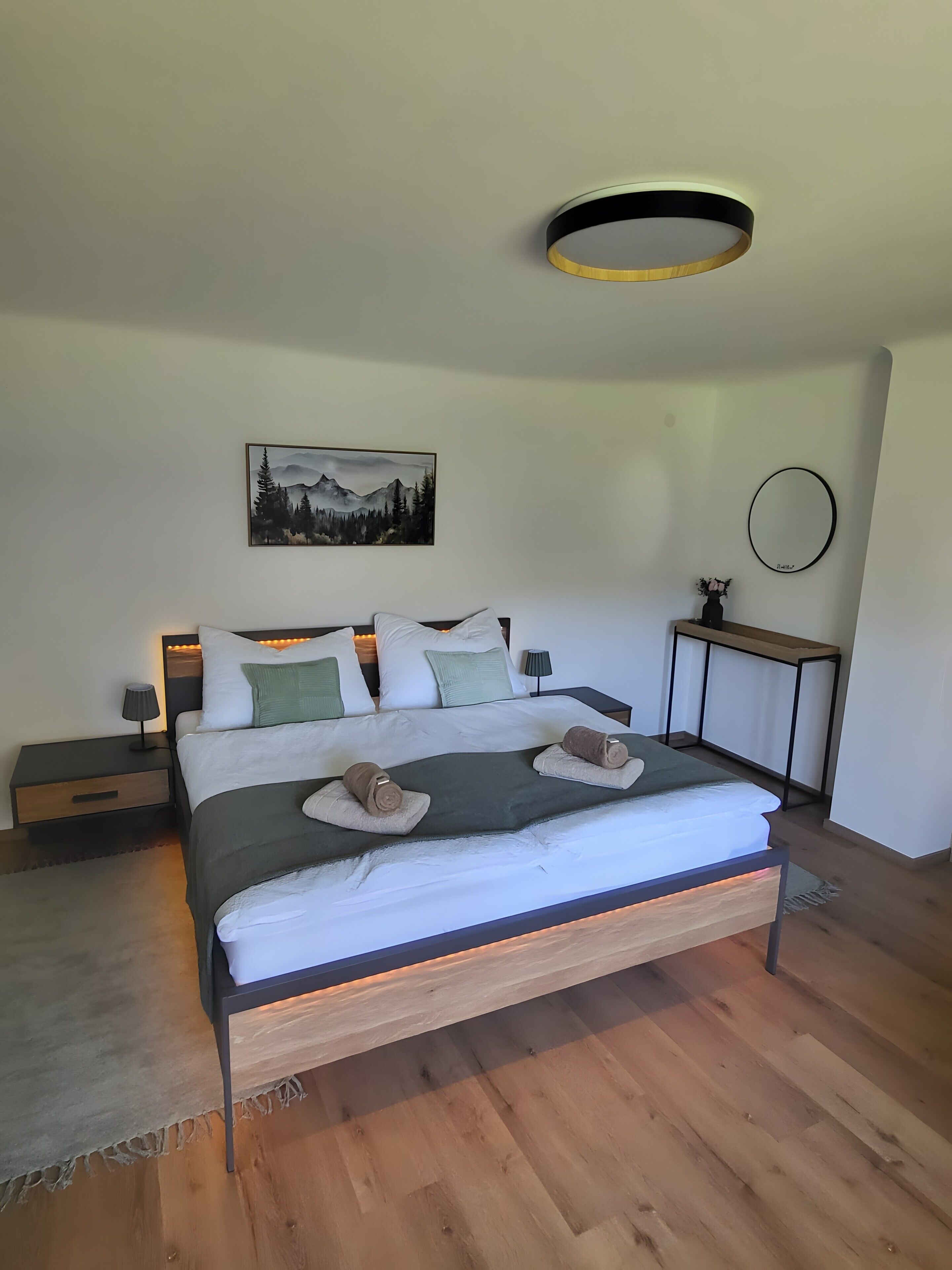 Ein modernes Schlafzimmer mit Holzboden, einem Bett mit weißer Bettwäsche, einem Nachttisch mit Lampe, einem an der Wand befestigten Spiegel und einem Gemälde von Bergen.