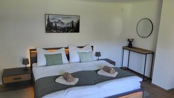 Ein modernes Schlafzimmer mit Holzboden, einem Bett mit weißer Bettwäsche, einem Nachttisch mit Lampe, einem an der Wand befestigten Spiegel und einem Gemälde von Bergen.