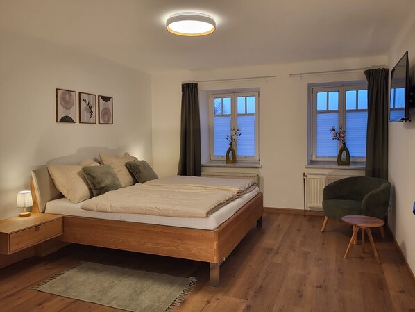 A bedroom with a wooden bed, bedside tables, a green armchair, and two windows with curtains.