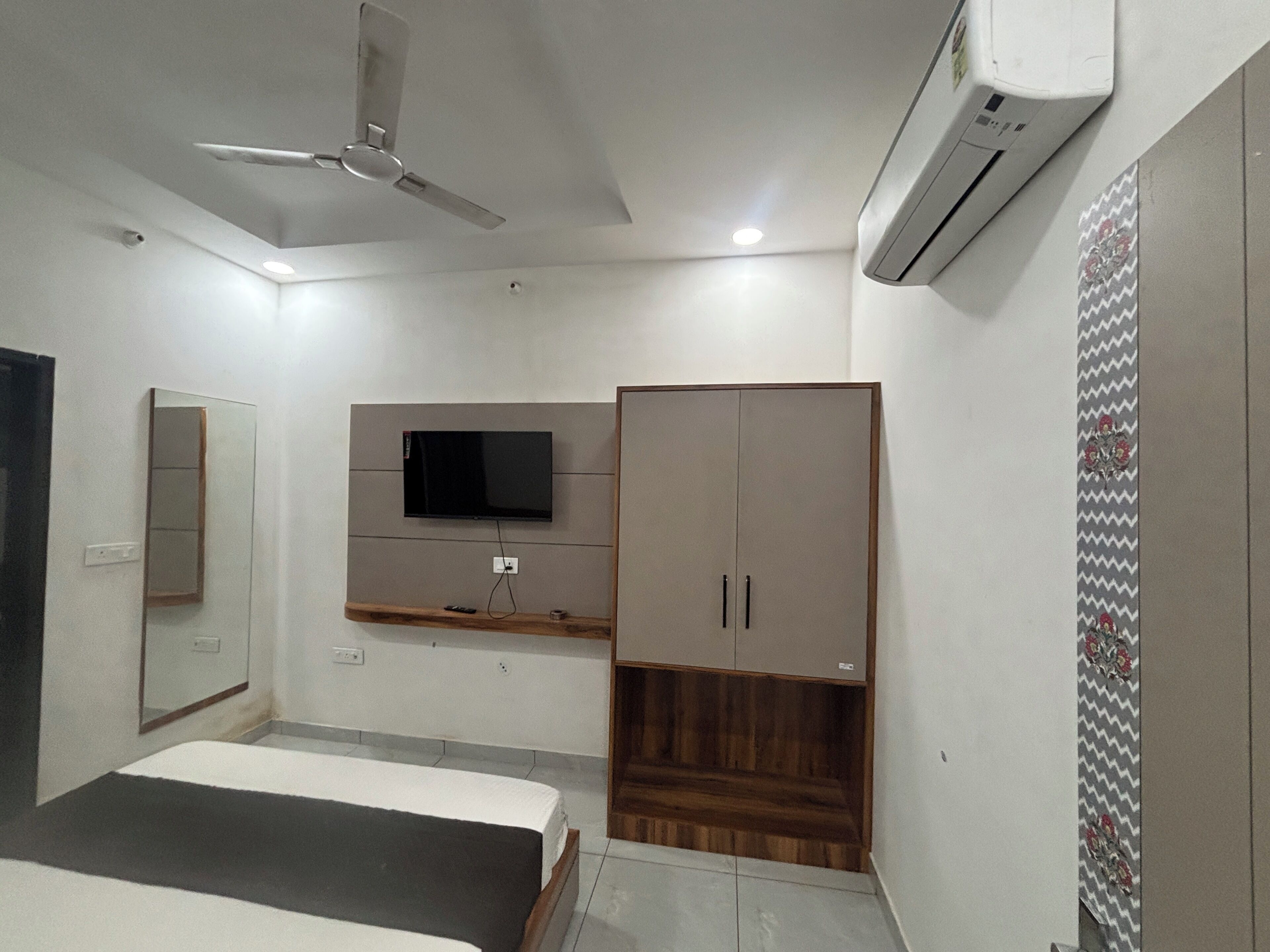 A bedroom with a bed, a TV mounted on the wall, a wardrobe, a ceiling fan, and an air conditioning unit.