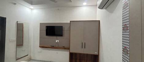 A bedroom with a bed, a TV mounted on the wall, a wardrobe, a ceiling fan, and an air conditioning unit.