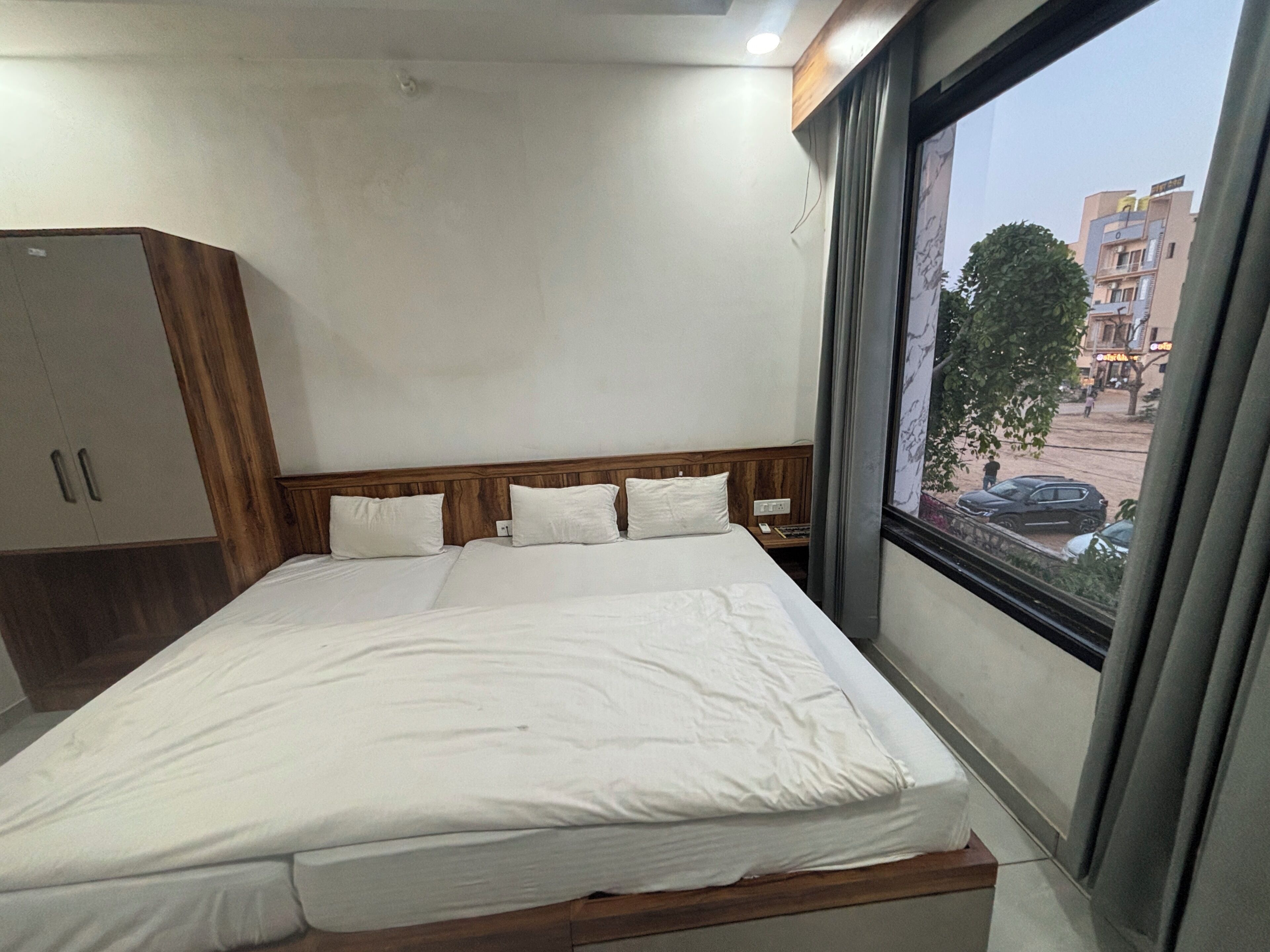 A hotel room with a bed, wooden headboard, white bedding, a wardrobe, and a window with a view of buildings and trees.