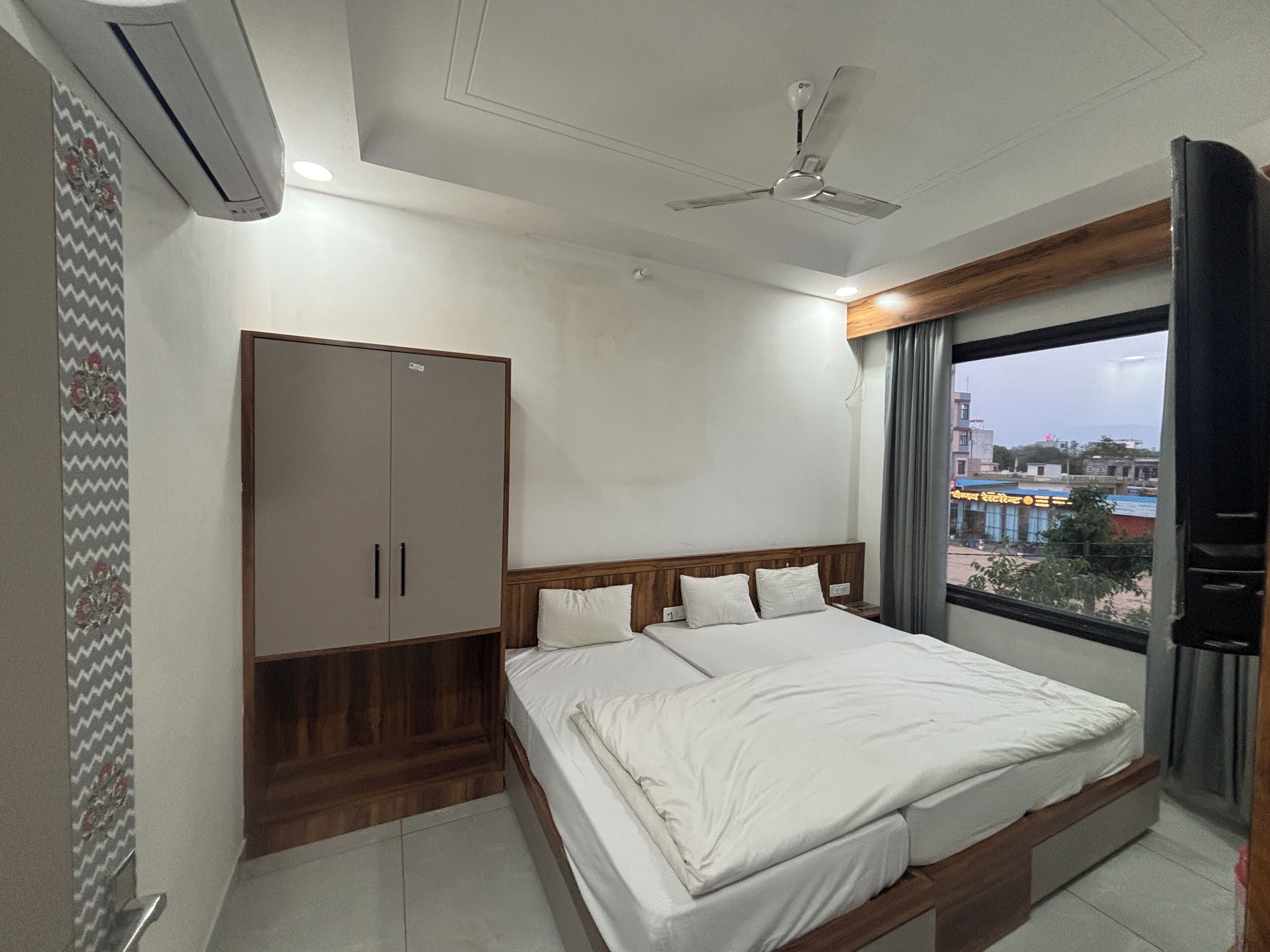 A bedroom with a bed, wardrobe, ceiling fan, and a window with a view of buildings.