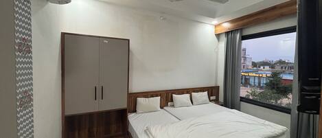 A bedroom with a bed, wardrobe, ceiling fan, and a window with a view of buildings.