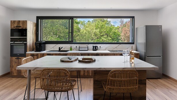 A modern kitchen with a large island, wooden cabinets, a built-in oven, and a view through a large window. - Loft Cavalier – Domaine de Greffeil: Unique Stay Overlooking the Equestrian Arena (Greffeil)