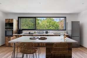 A modern kitchen with a large island, wooden cabinets, a built-in oven, and a view through a large window.