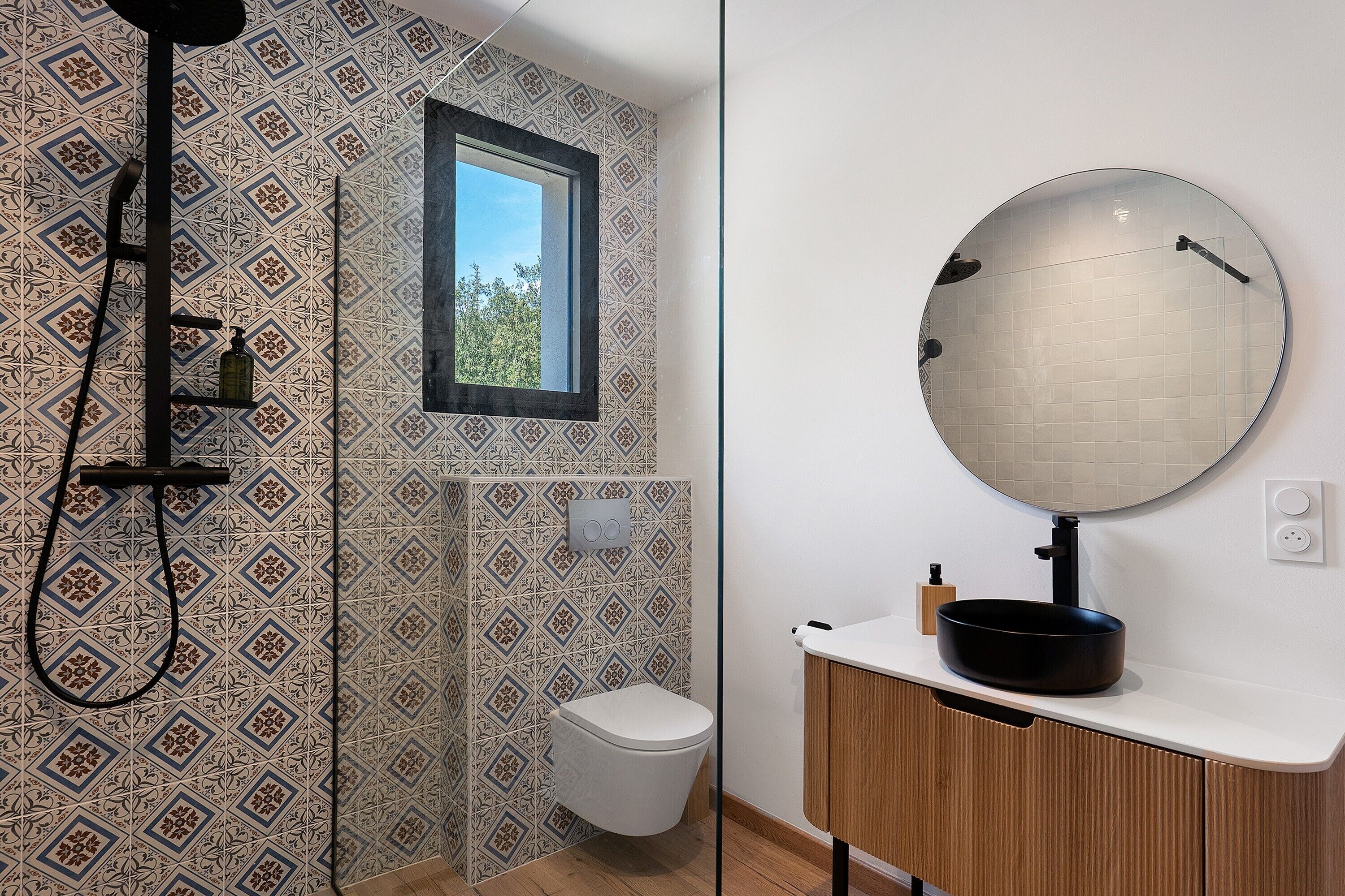 A bathroom with patterned tiles, a toilet, a shower with a black hose, a round mirror, and a white sink with a black basin.