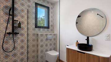A bathroom with patterned tiles, a toilet, a shower with a black hose, a round mirror, and a white sink with a black basin.