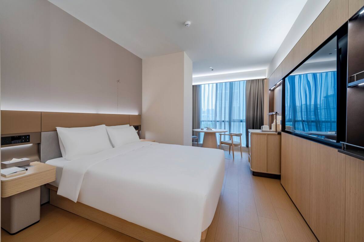 A modern hotel room with a large bed, a desk, a chair, a small table, and a large window with curtains.