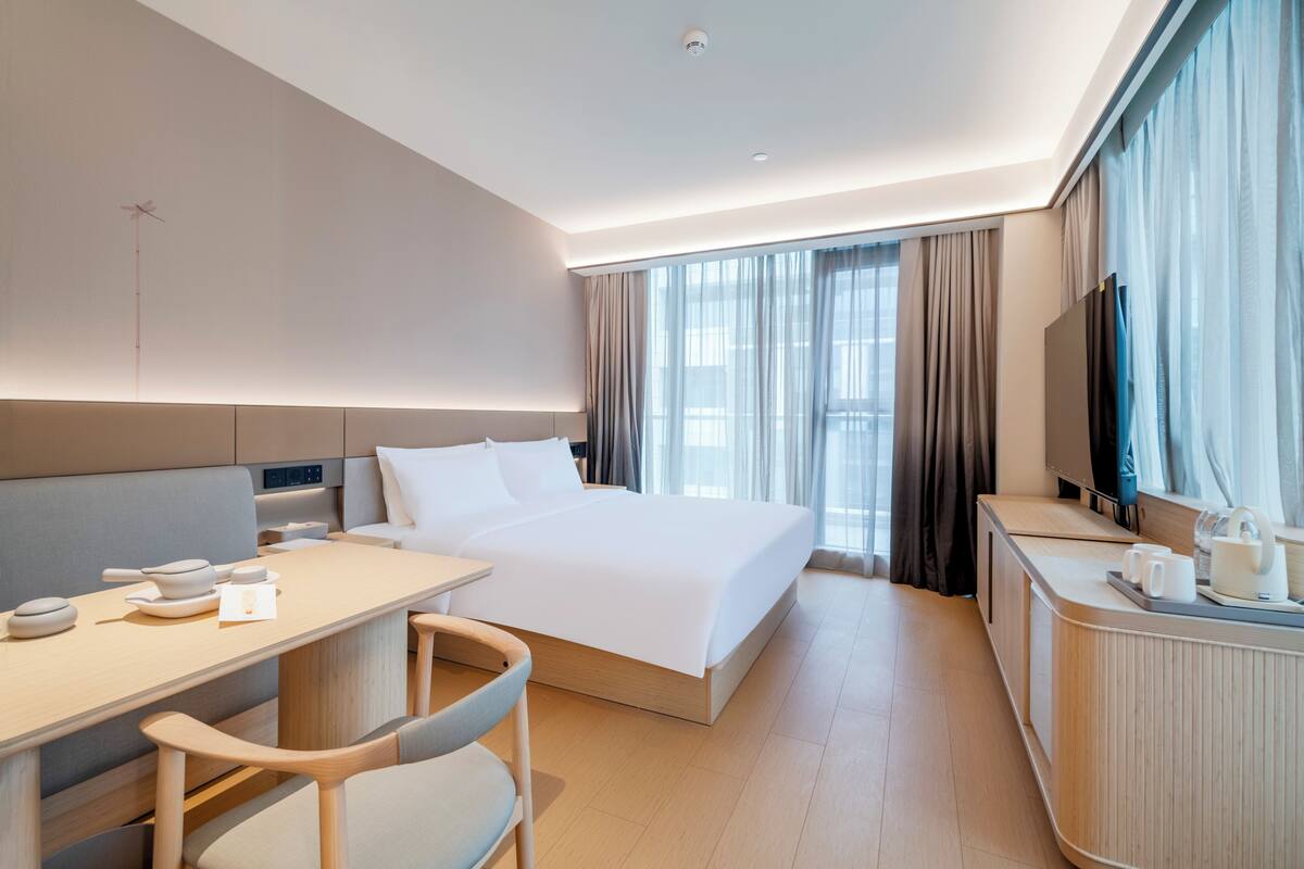 A modern hotel room with a bed, a desk, a chair, a television, and a window with curtains.