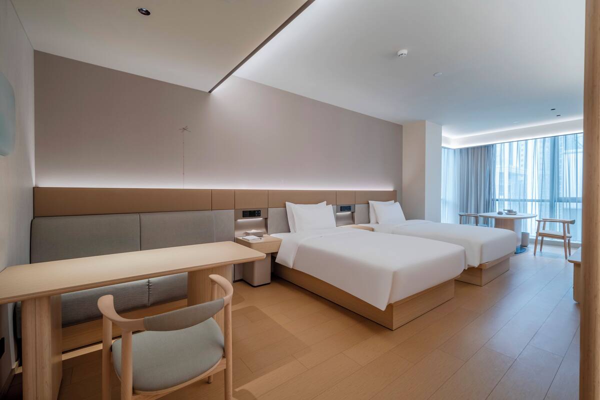 A modern hotel room with a large bed, a desk, a chair, and a view of the city through large windows.