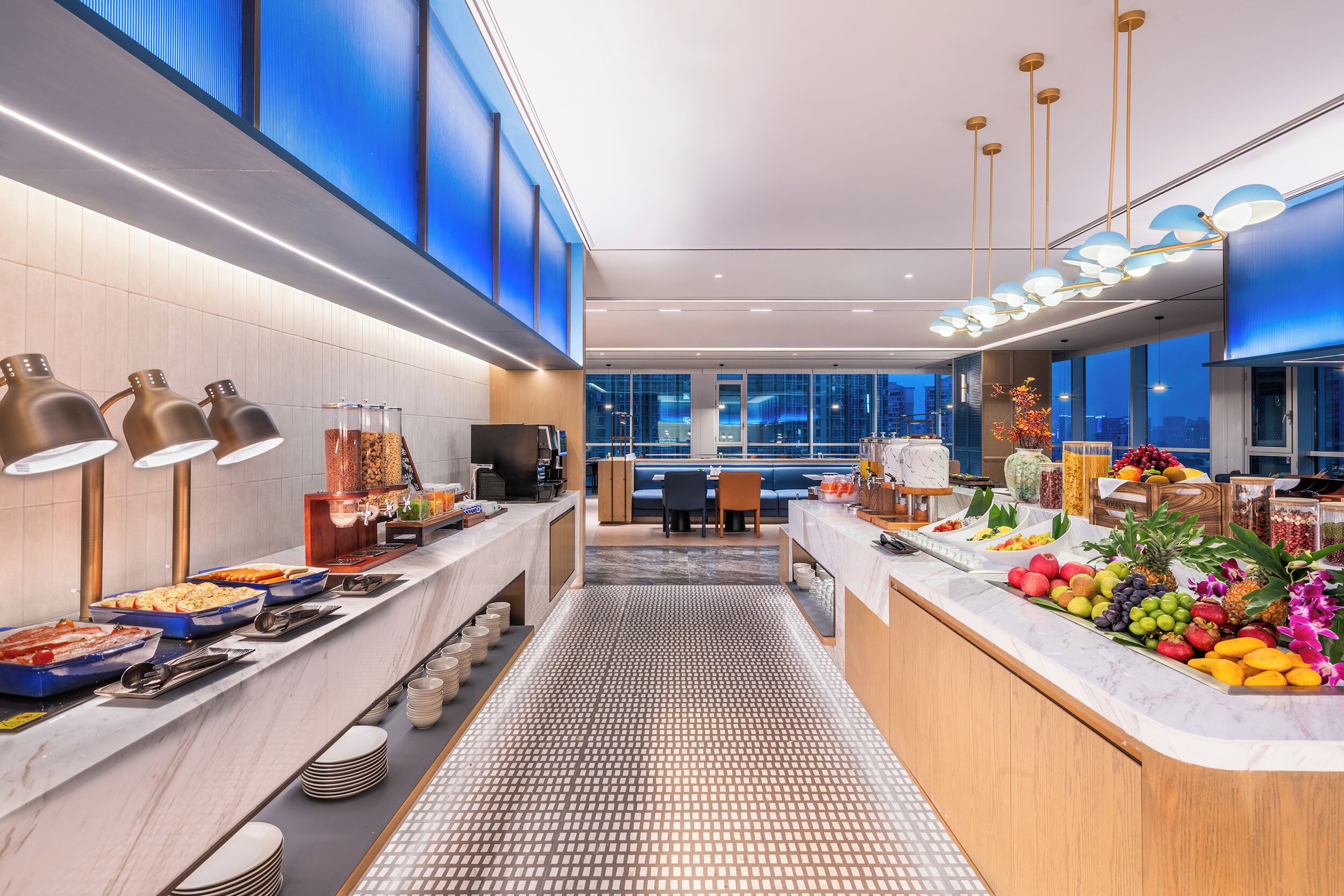 A modern hotel breakfast area with a buffet, seating area, and large windows.