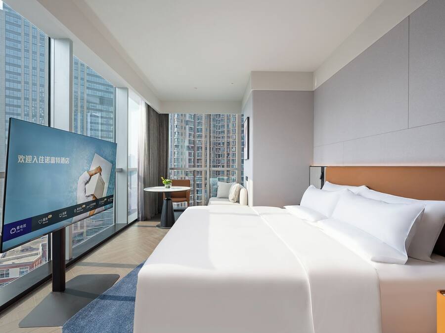 A modern hotel room with a large bed, a television, a small table with a chair, and a city view.