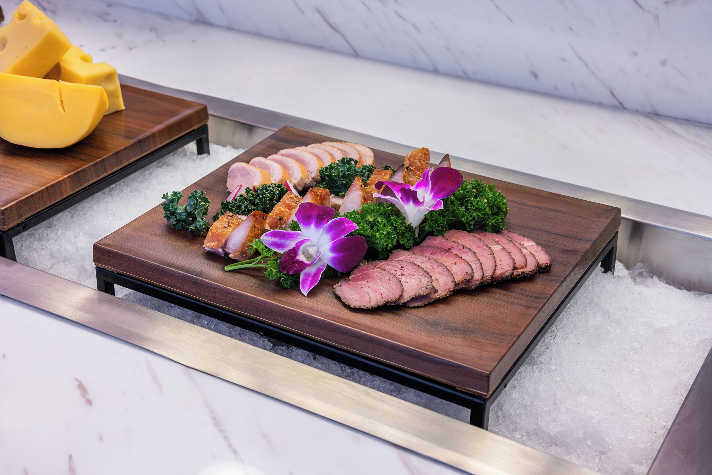 A wooden serving board with sliced meat, vegetables, and a yellow fruit on a marble surface.
