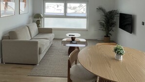 Ein modernes Wohnzimmer mit einer Couch, einem Esstisch, einem an der Wand befestigten Fernseher und einem großen Fenster.