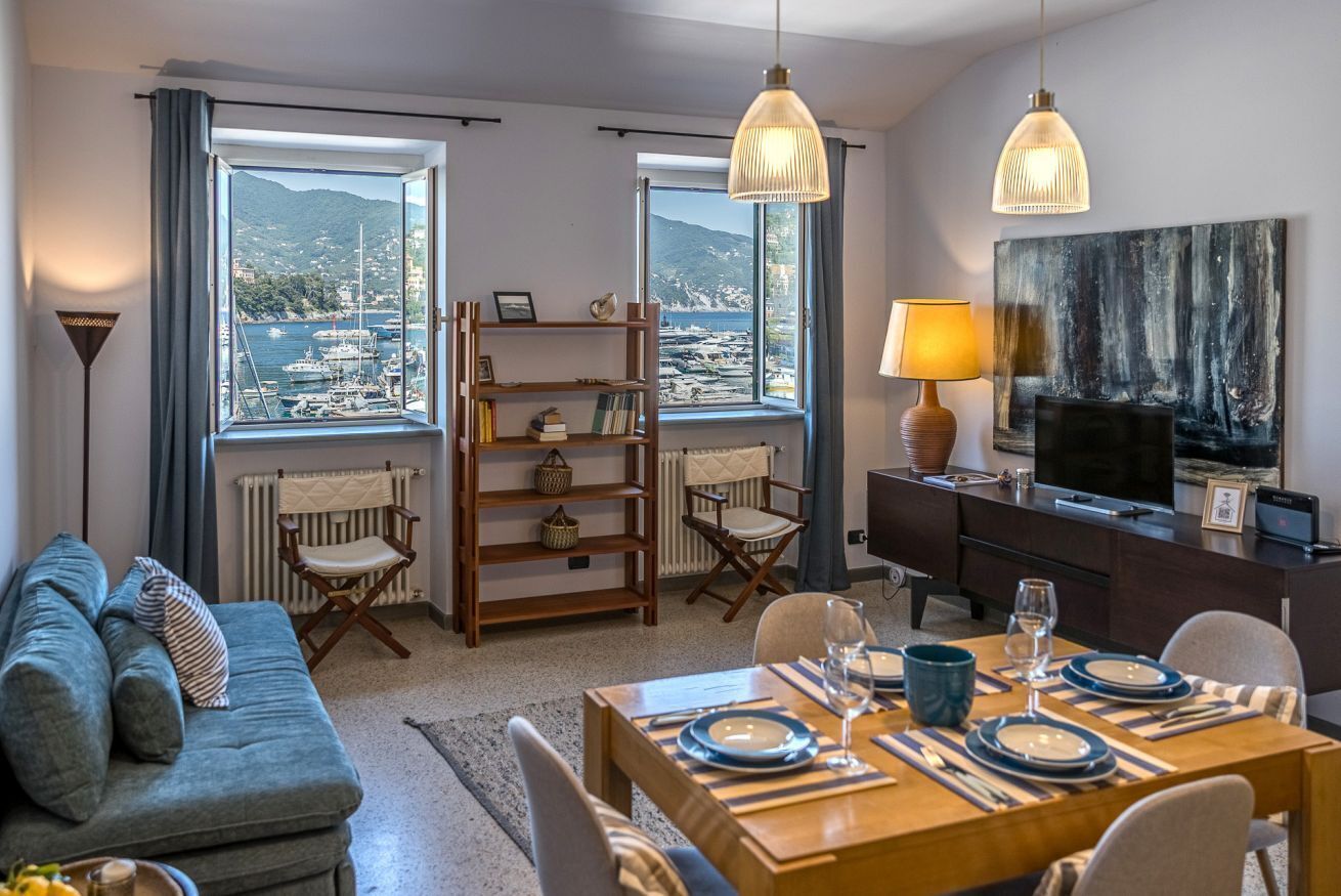 Un salon moderne comprenant un coin repas, de grandes fenêtres offrant une vue sur les bateaux et les collines, une bibliothèque et un téléviseur.