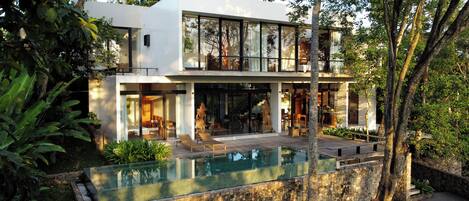 A modern two-story house with a glass facade, a swimming pool, and lush greenery.