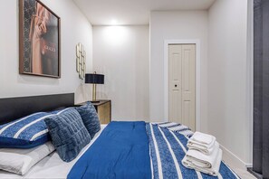 Un dormitorio con cama, almohadas, un cuadro enmarcado, una mesita de noche, una lámpara y una puerta.