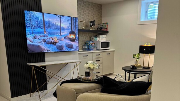 Ein modernes Wohnzimmer mit einem großen Flachbildfernseher, einem beigefarbenen Sofa, einem Glastisch und einer Küche im Hintergrund.