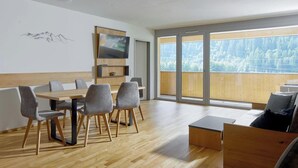 Una habitación moderna con una mesa de comedor de madera, sillas grises, un televisor de pantalla plana y un ventanal con vistas a árboles.