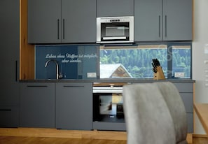 A modern kitchen with grey cabinets, a built-in oven, and a window with a view of a house. - Zapfig Living (Dalaas)