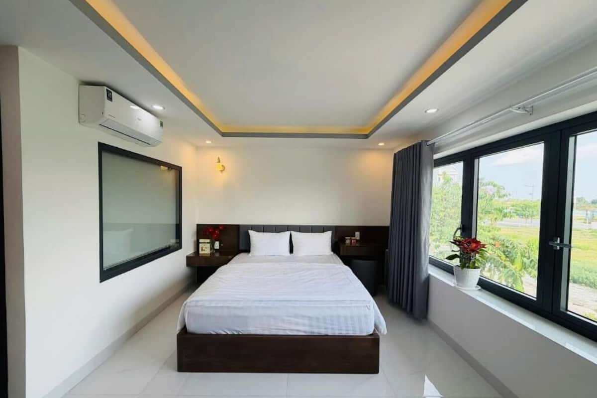 Una habitación de hotel moderna con una cama grande, una mesita de noche, aire acondicionado montado en la pared y una ventana con vistas a zonas verdes.