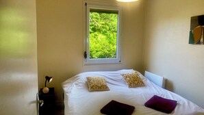 A bedroom with a bed, pillows, a nightstand, a lamp, a window with a view of greenery, and a hanging light fixture. - GITE DU MARAIS DE SUZANNE (Suzanne)