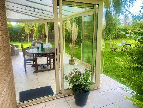 A glass door leading to an outdoor patio with a table and chairs, a potted plant, and a view of a lush garden.