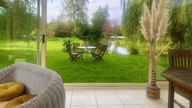 A patio with a table and chairs, a wicker chair, a potted plant, and a view of a pond and trees. - GITE DU MARAIS DE SUZANNE (Suzanne)