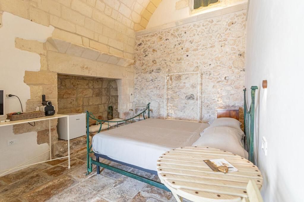 Una stanza con parete in pietra, un letto con struttura in metallo, una piccola scrivania con sedia e un camino.