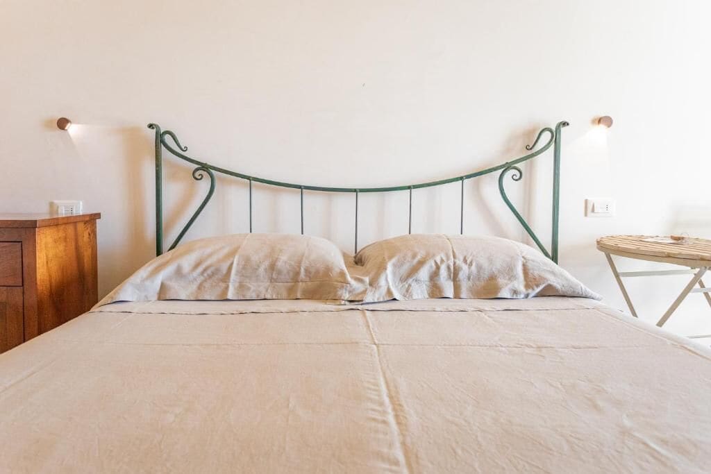 A bed with a curved metal headboard, two pillows, a wooden nightstand, and a small table with a chair.