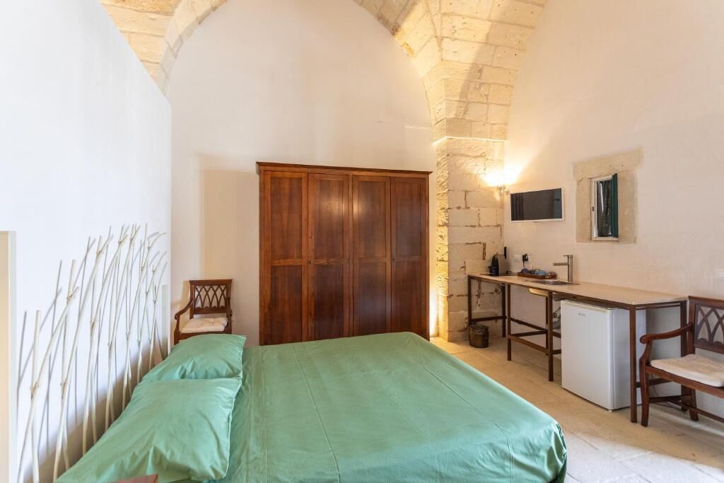 A bedroom with a bed, a wooden wardrobe, a desk with a chair, and a small kitchenette.