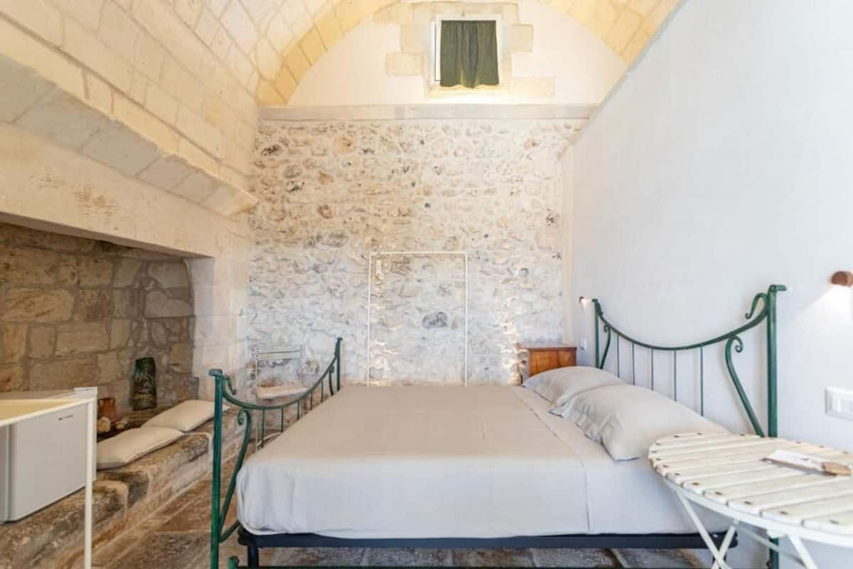 A stone-walled room with a bed, a small table, and a drying rack.