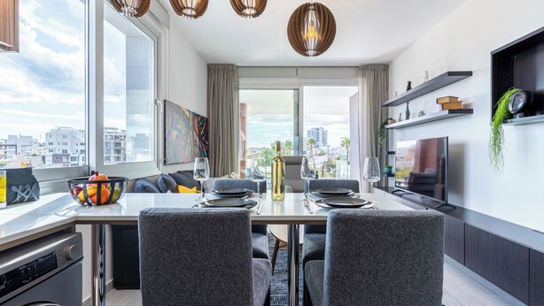 A modern dining area with a table set for four, a large window with a city view, and a kitchen area with a TV and shelves. - Sunflower 1-BR Apt. & Balcony in Larnaca (Larnaca)
