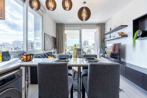 A modern dining area with a table set for four, a large window with a city view, and a kitchen area with a TV and shelves. - Sunflower 1-BR Apt. & Balcony in Larnaca (Larnaca)