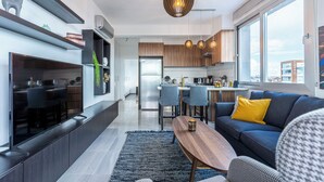 A modern living room with a sofa, coffee table, and a TV. There is a kitchen in the background with wooden cabinets and a dining area. - Sunflower 1-BR Apt. & Balcony in Larnaca (Larnaca)