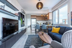 A modern living room with a sofa, coffee table, and a TV. There is a kitchen in the background with wooden cabinets and a dining area. - Sunflower 1-BR Apt. & Balcony in Larnaca (Larnaca)