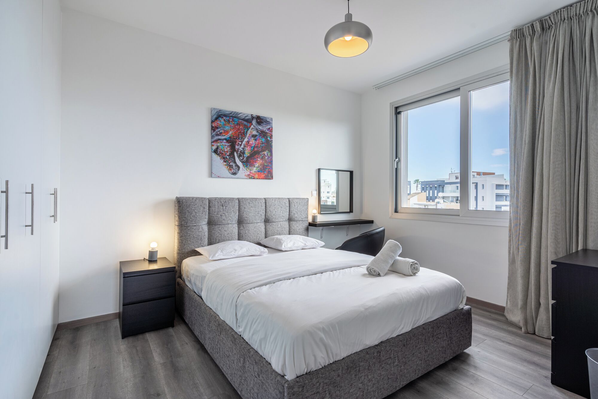 Ein modernes Hotelzimmer mit einem grau gepolsterten Bett, einem Nachttisch mit Lampe, einem großen Fenster mit Vorhängen und Blick auf die Stadt.