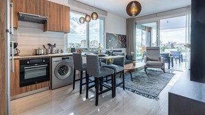 A modern kitchen with a built-in oven and washing machine, a dining area with a table and chairs, and a large window offering a city view. - Sunflower 1-BR Apt. & Balcony in Larnaca (Larnaca)