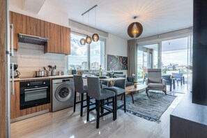 A modern kitchen with a built-in oven and washing machine, a dining area with a table and chairs, and a large window offering a city view. - Sunflower 1-BR Apt. & Balcony in Larnaca (Larnaca)