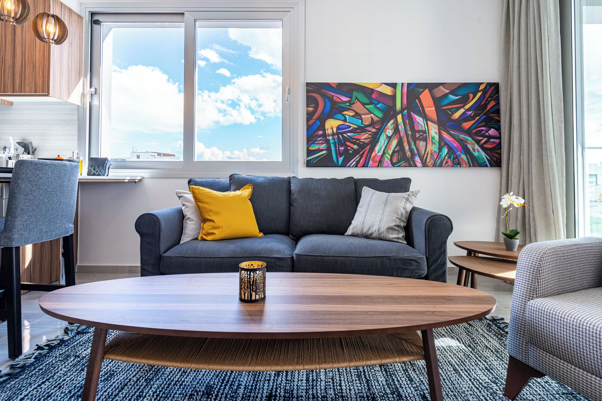 A modern living room with a sofa, a coffee table, a painting, and a view of the sky.