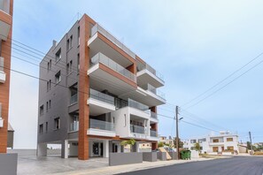 A multi-story residential building with balconies, a sign reading 'PLUS 27-B', and a clear sky. - Sunflower 1-BR Apt. & Balcony in Larnaca (Larnaca)