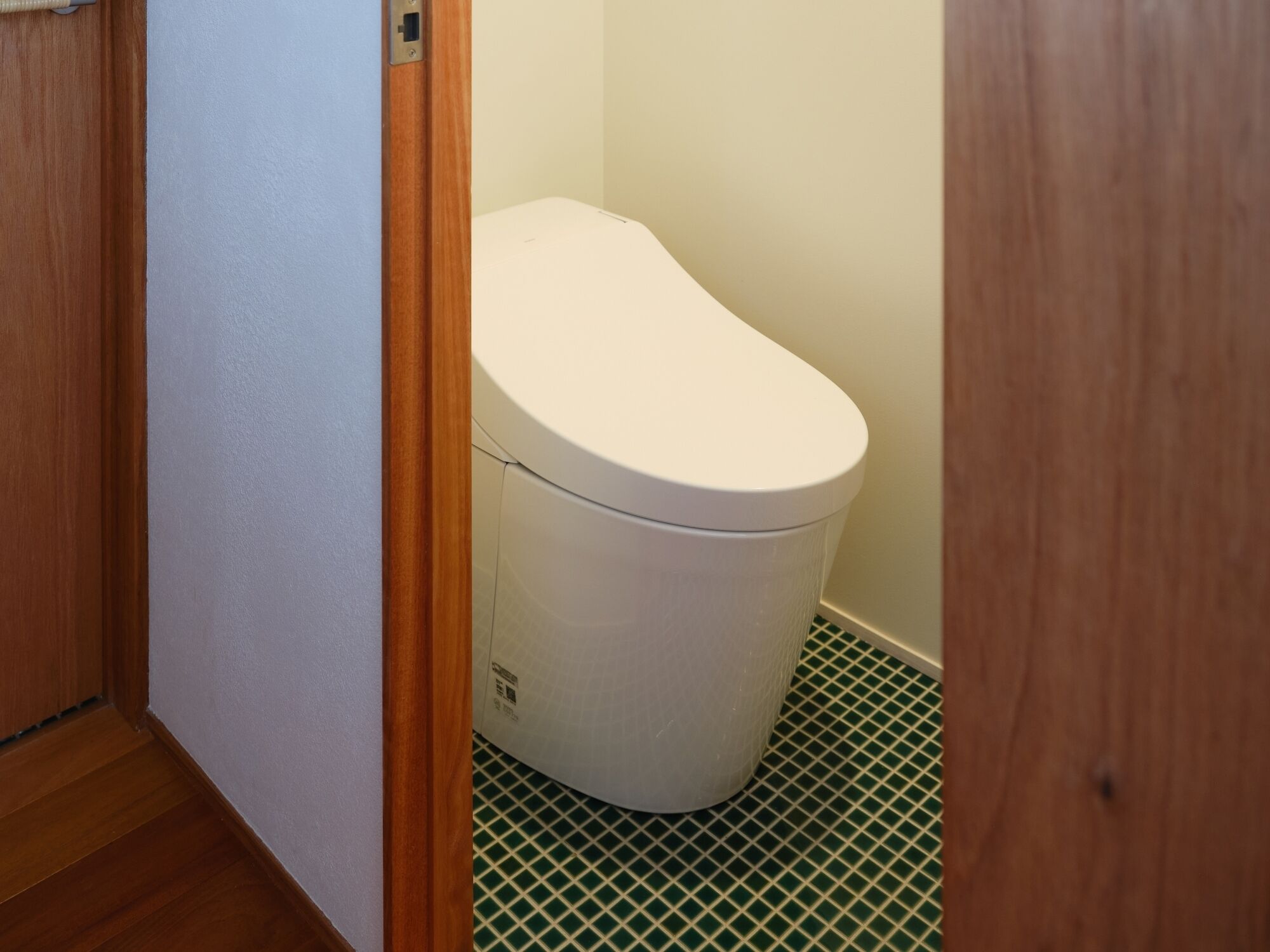 Ein kleines, schmales Badezimmer mit einer weißen Toilette, einem hölzernen Türrahmen und einem grün gefliesten Boden.