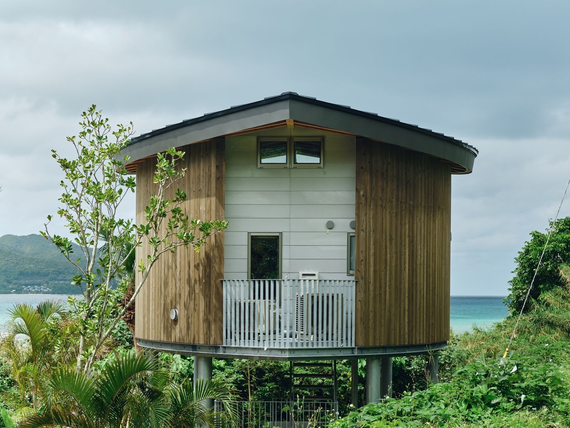 A modern, elevated wooden house with a balcony, situated by a body of water with mountains in the background.