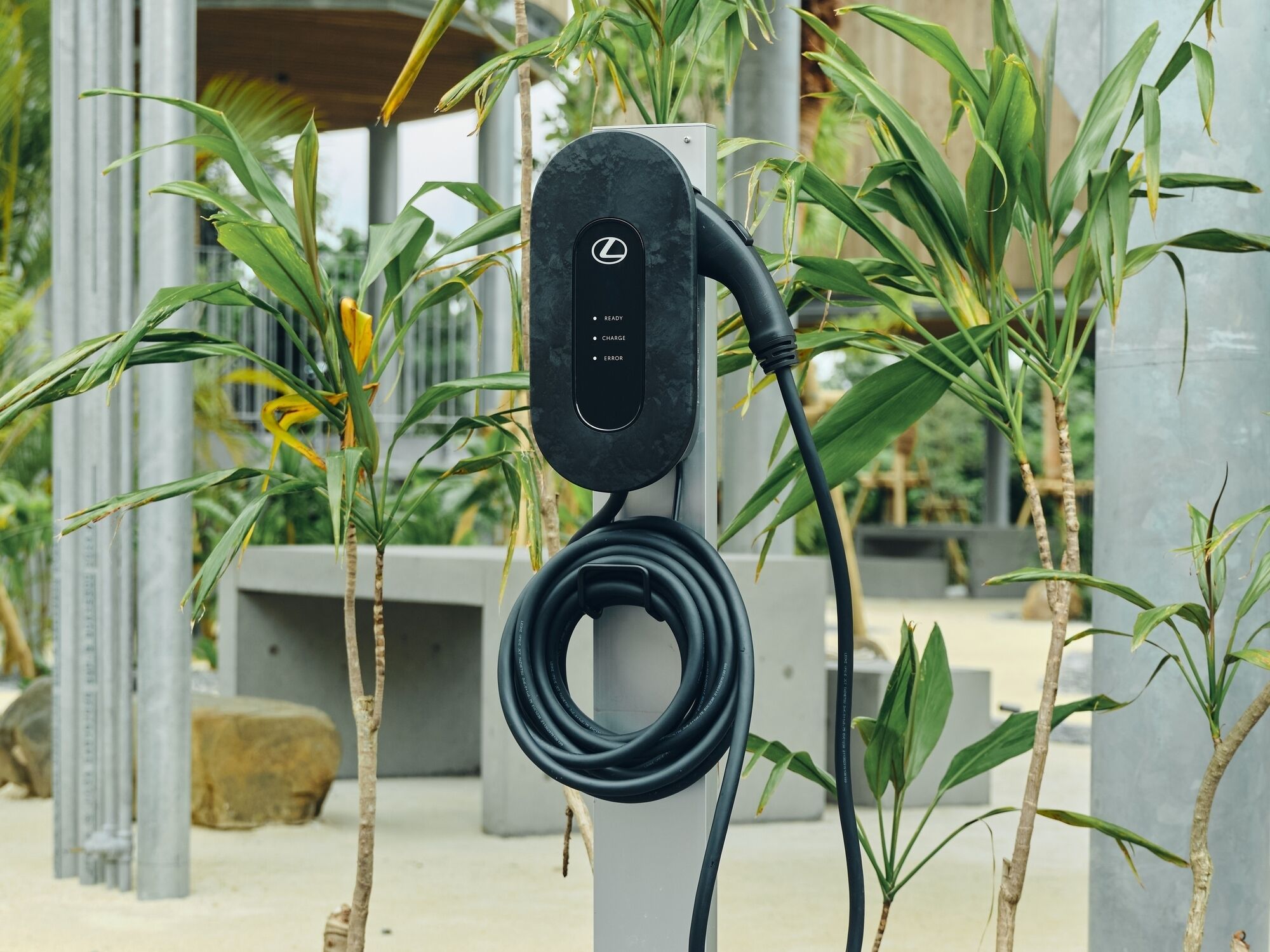 A black electric vehicle charging station with a cable, surrounded by greenery and architectural columns.