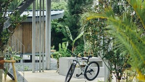 Ein Fahrrad, das an einer Betonkonstruktion in einem Garten lehnt, mit Pflanzen und einem Holzgebäude im Hintergrund.